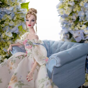 May include: A porcelain doll with blonde hair and a floral dress sits on a blue velvet sofa. The doll is surrounded by blue and white flowers.