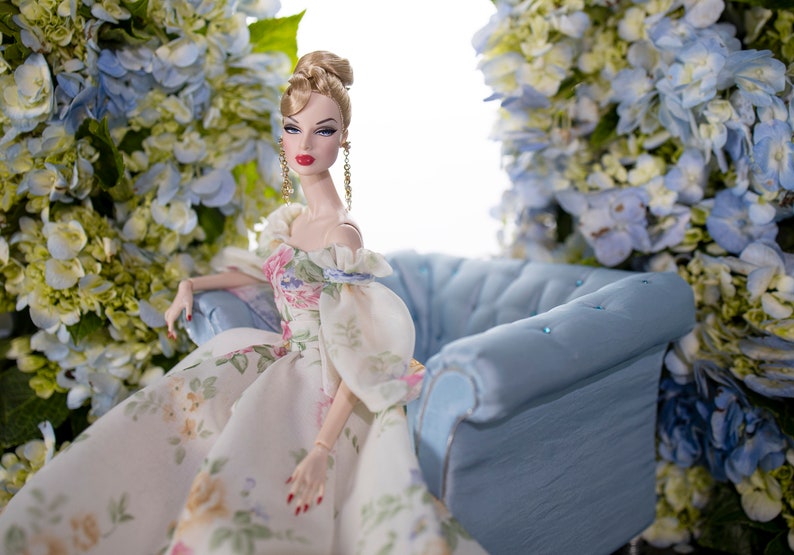 May include: A porcelain doll with blonde hair and a floral dress sits on a blue velvet sofa. The doll is surrounded by blue and white flowers.