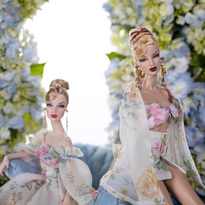 May include: Two fashion dolls wearing floral print dresses. The doll on the left is sitting on a blue chair. The doll on the right is standing with her arms at her sides. Both dolls have blonde hair and are wearing floral print dresses. The background is a wall of blue and white flowers.