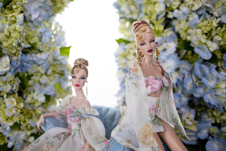 May include: Two fashion dolls wearing floral print dresses. The doll on the left is sitting on a blue chair. The doll on the right is standing with her arms at her sides. Both dolls have blonde hair and are wearing floral print dresses. The background is a wall of blue and white flowers.