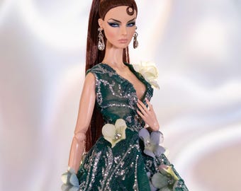 May include: A fashion doll wearing a green sequined gown with floral embellishments. The doll has long brown hair, blue eyes, and is wearing a blue bow in her hair.