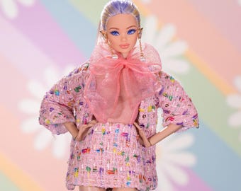 Puede incluir: Una muñeca rosa y blanca que lleva una chaqueta y una falda de tweed rosa con un gran lazo rosa. La muñeca tiene el pelo azul y morado y los ojos azules.
