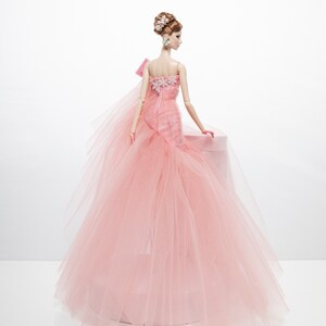 May include: A pink tulle ball gown with a floral bodice and a bow on the shoulder. The doll is wearing the gown and has her hair styled in an updo with flowers.