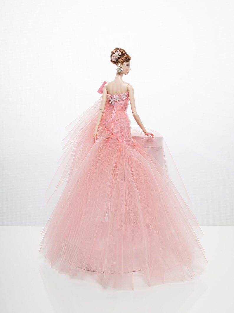 May include: A pink tulle ball gown with a floral bodice and a bow on the shoulder. The doll is wearing the gown and has her hair styled in an updo with flowers.