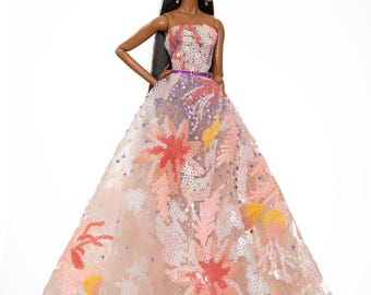 May include: A fashion doll wearing a strapless gown with a floral pattern and sequins. The gown is pink, orange, and yellow with a purple belt. The doll has long black hair and brown eyes.