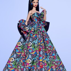 May include: A fashion doll wearing a strapless gown with a colorful, patterned design. The dress features a large black bow in the back and a matching bow on the left shoulder. The doll has long black hair, red lipstick, and blue earrings.