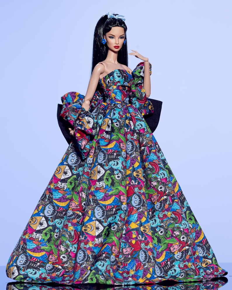 May include: A fashion doll wearing a strapless gown with a colorful, patterned design. The dress features a large black bow in the back and a matching bow on the left shoulder. The doll has long black hair, red lipstick, and blue earrings.
