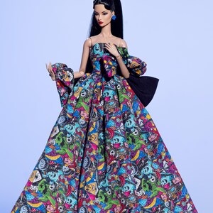 May include: A fashion doll wearing a colorful, off-the-shoulder gown. The dress features a vibrant print with skulls and other cartoonish figures in shades of blue, green, pink, and yellow. The doll has long black hair, blue earrings, and a matching hair accessory.