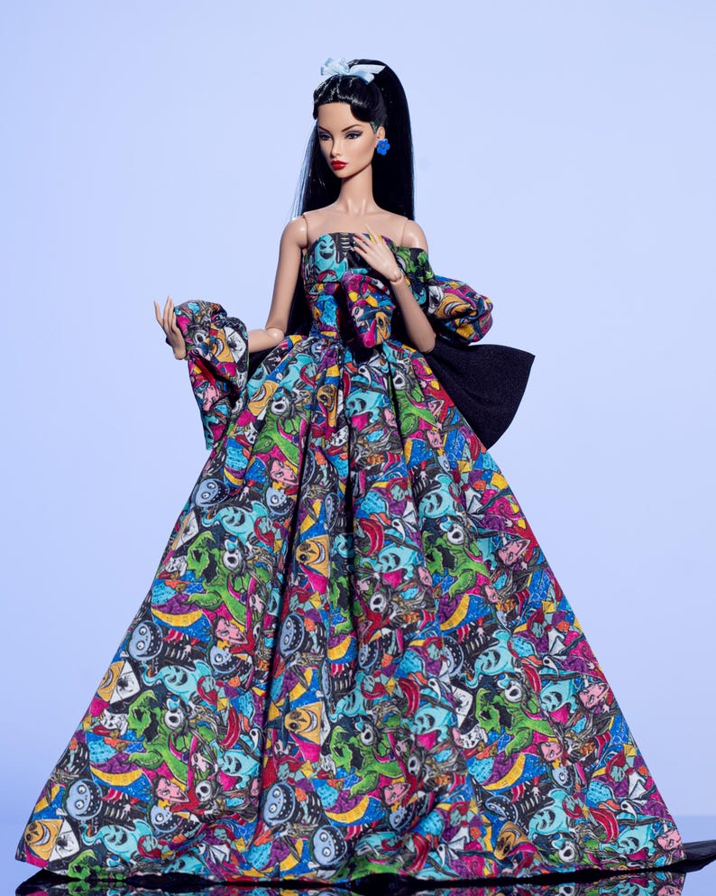 May include: A fashion doll wearing a colorful, off-the-shoulder gown. The dress features a vibrant print with skulls and other cartoonish figures in shades of blue, green, pink, and yellow. The doll has long black hair, blue earrings, and a matching hair accessory.