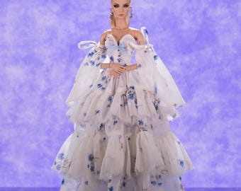 May include: A porcelain doll wearing a white dress with blue floral print and a blue bow. The dress has a layered, ruffled skirt and off-the-shoulder sleeves.