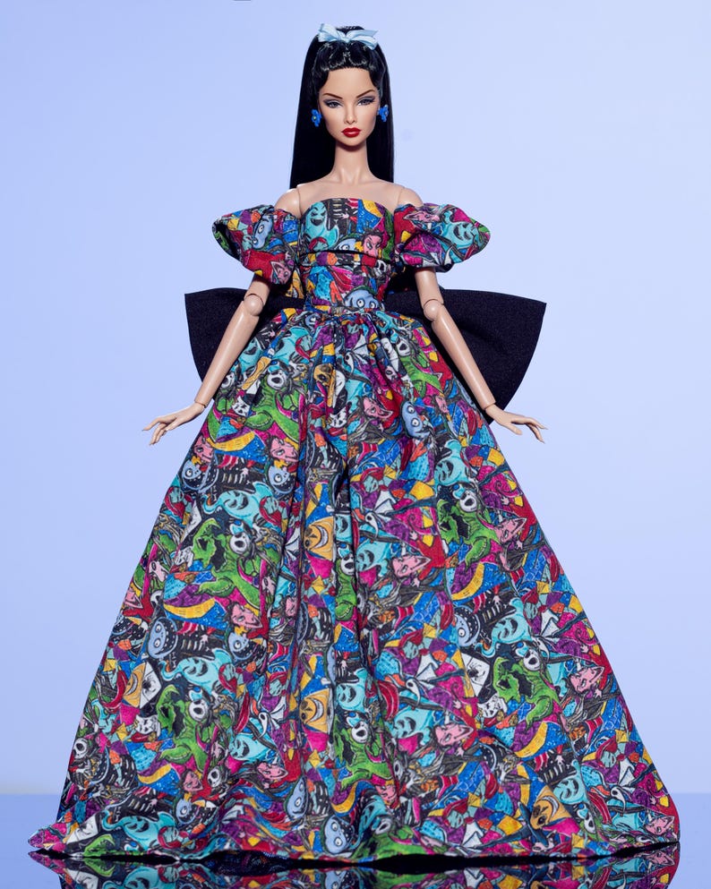 May include: A fashion doll wearing a colorful, off-the-shoulder gown with a black bow. The dress features a vibrant print with cartoon-like characters in various colors. The doll has long black hair, blue earrings, and a matching headband.