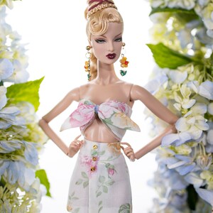 May include: A doll with blonde hair styled in a bun with braids, wearing a white floral print top and matching skirt. The doll is posed with her arms crossed and is surrounded by blue and white flowers.