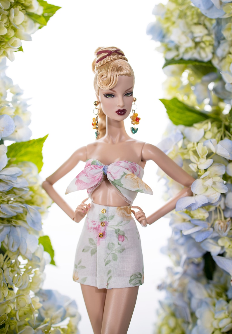 May include: A doll with blonde hair styled in a bun with braids, wearing a white floral print top and matching skirt. The doll is posed with her arms crossed and is surrounded by blue and white flowers.