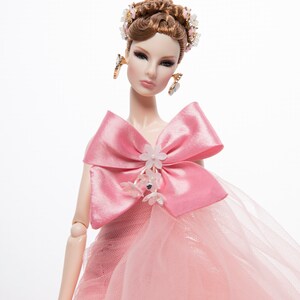 May include: A pink doll with brown hair styled in an updo with a floral hairpiece. The doll is wearing a pink tulle gown with a large pink satin bow and floral embellishments.
