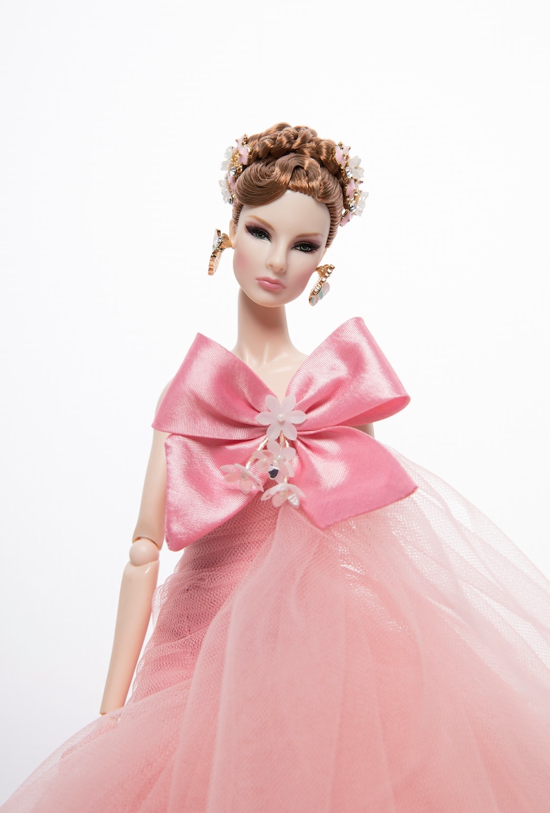 May include: A pink doll with brown hair styled in an updo with a floral hairpiece. The doll is wearing a pink tulle gown with a large pink satin bow and floral embellishments.