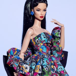 May include: A fashion doll with long black hair, red lipstick, and blue flower earrings. The doll wears a strapless dress with a colorful, cartoon-like print and a large black bow. A light blue bow adorns the doll's hair.