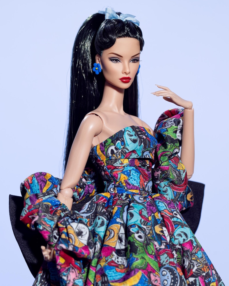 May include: A fashion doll with long black hair, red lipstick, and blue flower earrings. The doll wears a strapless dress with a colorful, cartoon-like print and a large black bow. A light blue bow adorns the doll's hair.