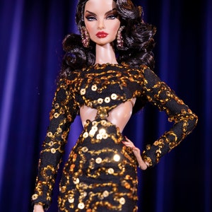 Puede incluir: Una mu&ntilde;eca de moda con el pelo largo y negro y un vestido de lentejuelas negras y doradas. La mu&ntilde;eca lleva pendientes grandes y tiene un maquillaje dram&aacute;tico.