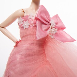 May include: A pink tulle gown with a large pink satin bow and white flower embellishments. The gown has a strapless neckline and a fitted bodice.