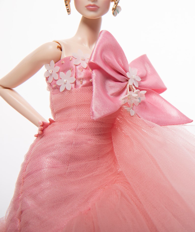 May include: A pink tulle gown with a large pink satin bow and white flower embellishments. The gown has a strapless neckline and a fitted bodice.