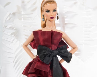 May include: A fashion doll wearing a red satin jumpsuit with a large black bow. The doll has blonde hair pulled back in a ponytail and is wearing black earrings.