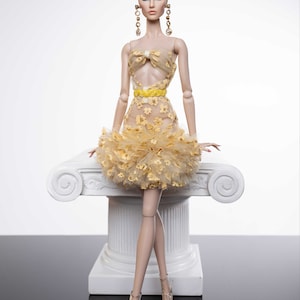 May include: A fashion doll wearing a yellow and white floral dress with a yellow bow and a ruffled skirt. The doll is sitting on a white pedestal with a scroll design.