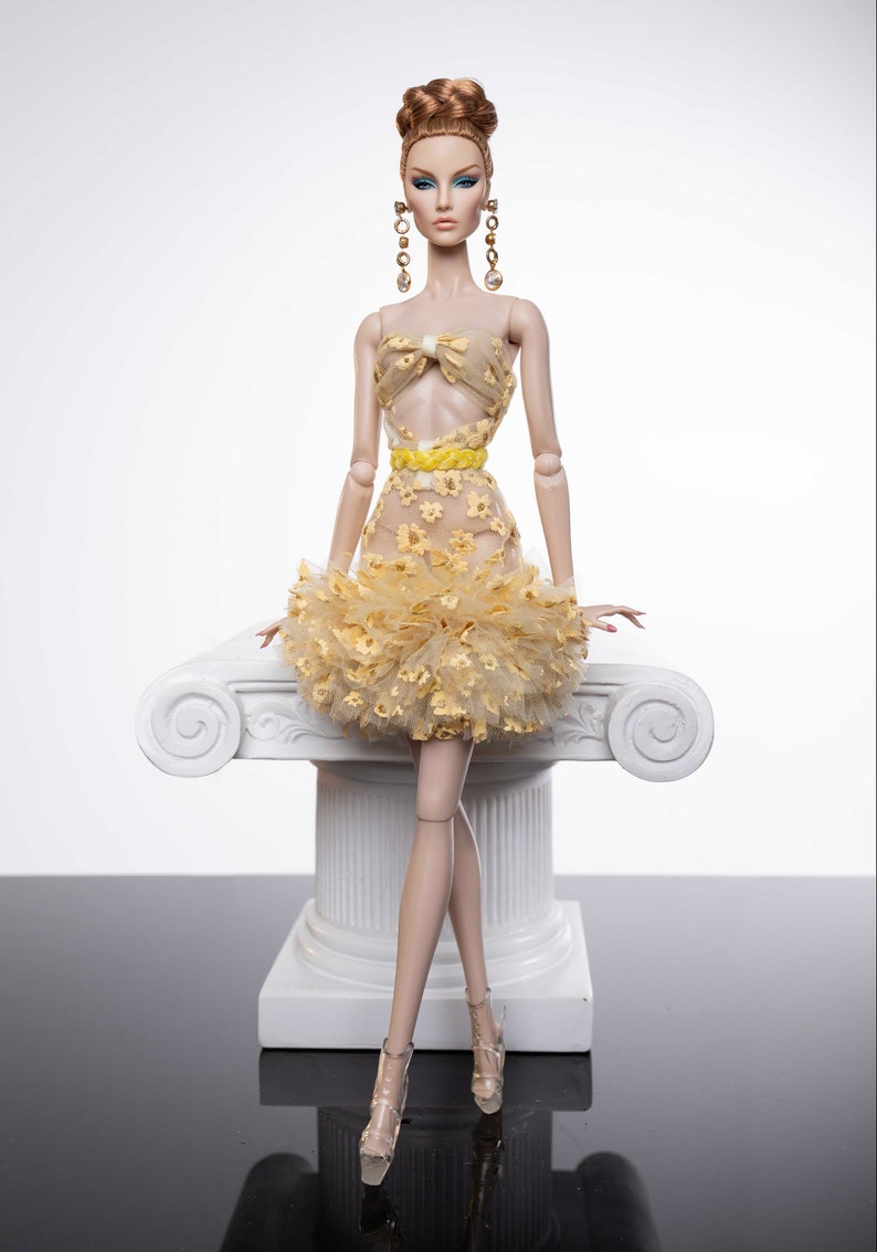 May include: A fashion doll wearing a yellow and white floral dress with a yellow bow and a ruffled skirt. The doll is sitting on a white pedestal with a scroll design.