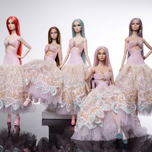 May include: Five fashion dolls with different hair colors, including red, brown, and blue, are posed in a studio setting. Each doll wears a light pink dress with a layered, lacy skirt, embellished with gold and turquoise accents. The dolls are standing on a reflective surface.