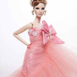 May include: A pink tulle ball gown with a large pink satin bow and white flower embellishments. The doll is wearing a gold crown and earrings.