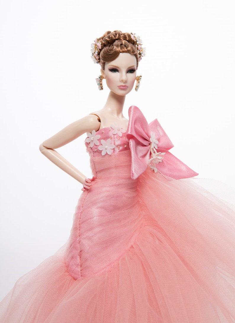 May include: A pink tulle ball gown with a large pink satin bow and white flower embellishments. The doll is wearing a gold crown and earrings.