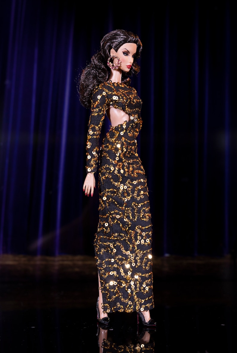 Puede incluir: Una mu&ntilde;eca de moda con un vestido de lentejuelas negro y dorado con un detalle de corte. La mu&ntilde;eca tiene el pelo oscuro y lleva grandes pendientes de aro.