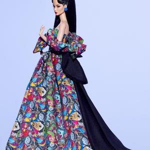 May include: A fashion doll wearing a colorful, patterned gown with a black train. The gown features a variety of cartoon faces in vibrant colors. The doll has long black hair, red lipstick, and blue earrings. A white bow adorns the doll's hair.
