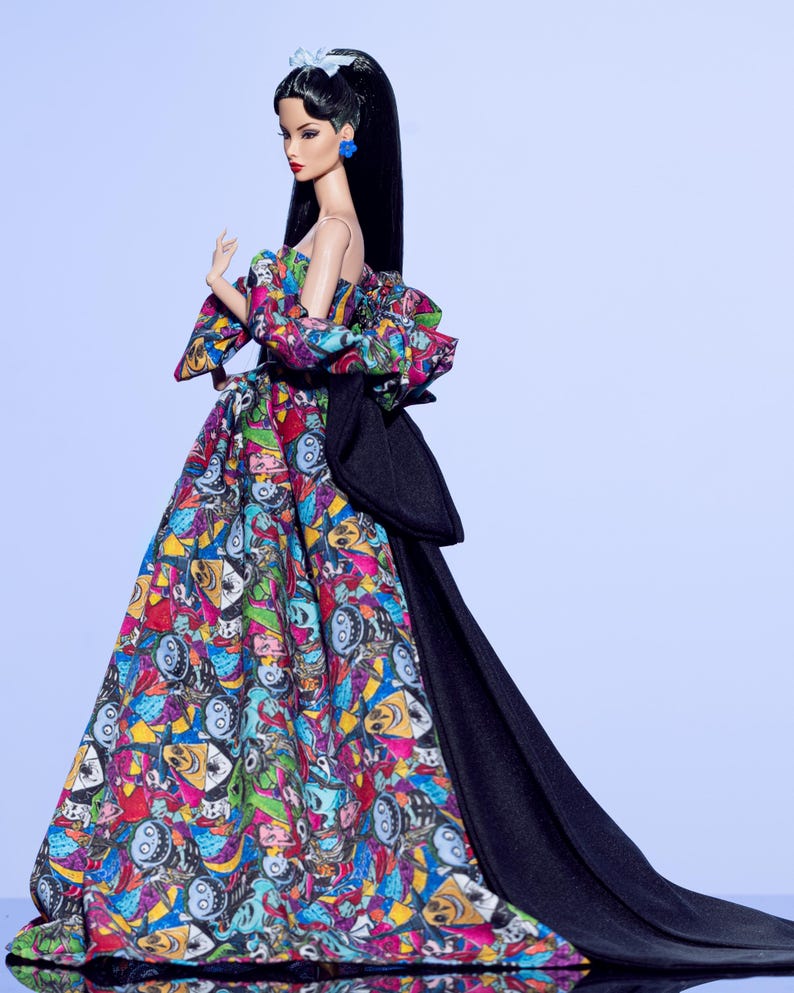 May include: A fashion doll wearing a colorful, patterned gown with a black train. The gown features a variety of cartoon faces in vibrant colors. The doll has long black hair, red lipstick, and blue earrings. A white bow adorns the doll's hair.