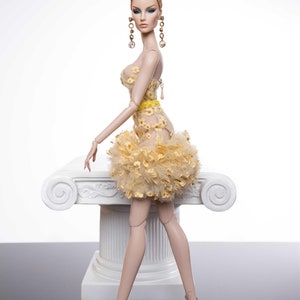 May include: A fashion doll with long, auburn hair styled in a bun, wearing a yellow and gold dress with a ruffled skirt and gold earrings. The doll is standing on a white pedestal with a decorative scroll design.