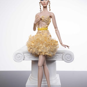 May include: A fashion doll wearing a yellow and white dress with a ruffled skirt. The doll has long brown hair styled in a bun and is wearing gold earrings. The doll is sitting on a white pedestal.