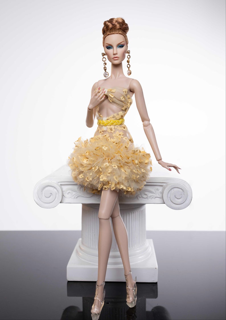May include: A fashion doll wearing a yellow and white dress with a ruffled skirt. The doll has long brown hair styled in a bun and is wearing gold earrings. The doll is sitting on a white pedestal.