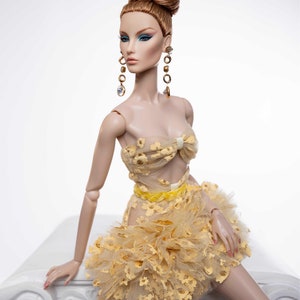 May include: A fashion doll wearing a yellow and gold dress with a ruffled skirt and a bow at the top. The doll has long, red hair styled in a bun and is wearing gold earrings with clear stones.