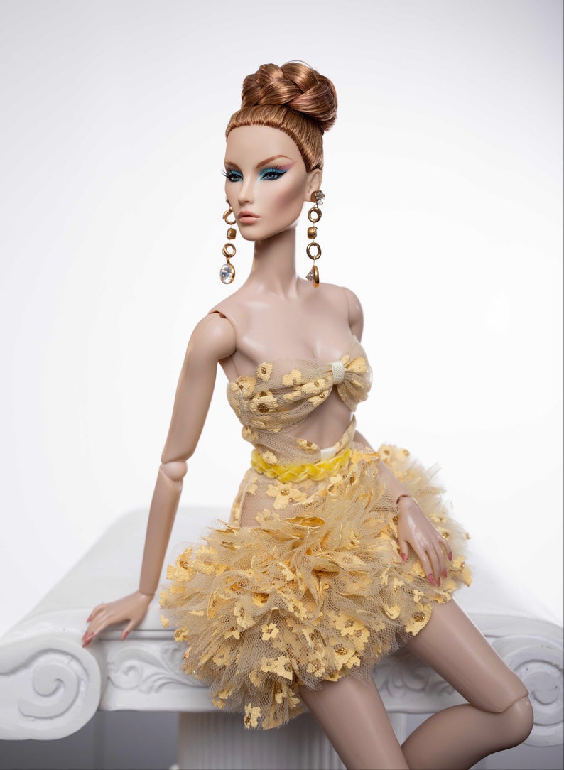 May include: A fashion doll wearing a yellow and gold dress with a ruffled skirt and a bow at the top. The doll has long, red hair styled in a bun and is wearing gold earrings with clear stones.