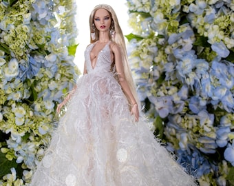 May include: A fashion doll wearing a white, fluffy gown with a plunging neckline. The doll has long blonde hair and is standing in front of a backdrop of blue and white flowers.