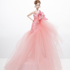 May include: A pink tulle ball gown with a floral design and a large pink bow. The doll is wearing the gown and has brown hair and a light pink blush.
