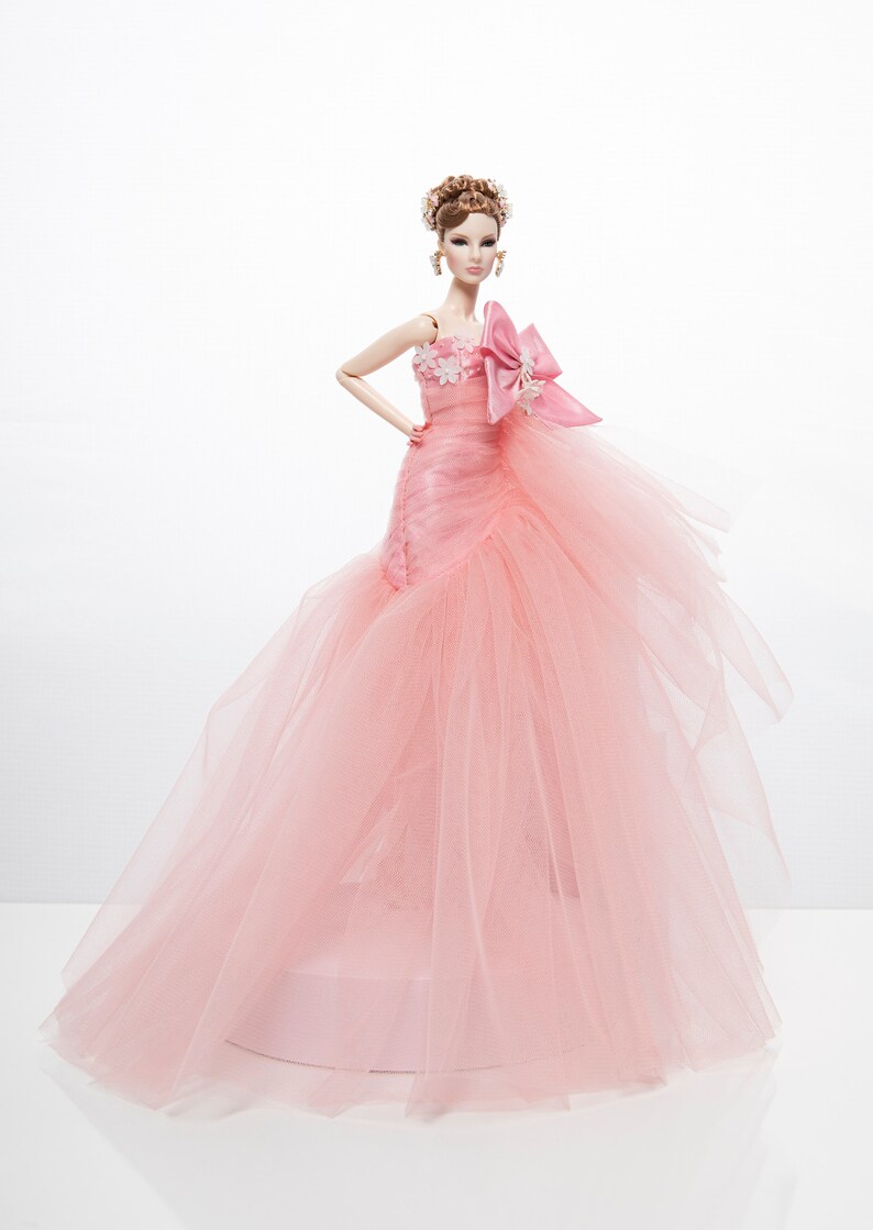 May include: A pink tulle ball gown with a floral design and a large pink bow. The doll is wearing the gown and has brown hair and a light pink blush.