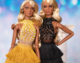 Puede incluir: Dos muñecas de moda con cabello largo rubio que llevan tops halter con cuentas y faldas de flecos en capas. Una muñeca lleva una falda negra y la otra una falda amarilla.