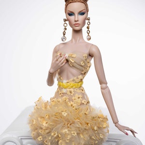May include: A fashion doll wearing a yellow and white floral dress with a yellow belt. The doll has long brown hair styled in a bun and is wearing gold earrings with clear stones.
