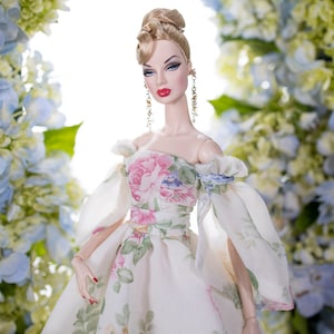 May include: A porcelain doll wearing a white dress with a floral print. The doll has blonde hair styled in an updo and is wearing gold earrings. The doll is posed against a backdrop of blue and white flowers.