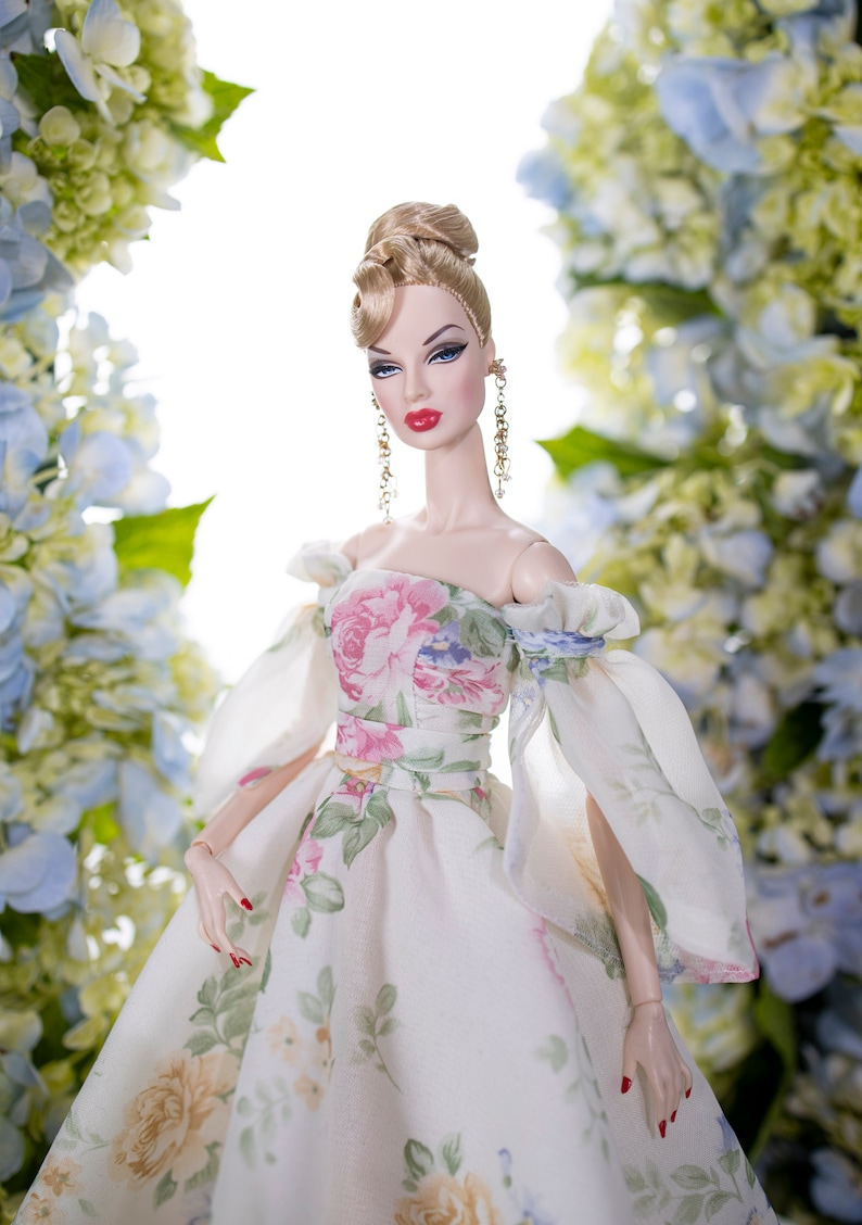 May include: A porcelain doll wearing a white dress with a floral print. The doll has blonde hair styled in an updo and is wearing gold earrings. The doll is posed against a backdrop of blue and white flowers.