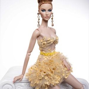 May include: A fashion doll with long, auburn hair styled in a bun, wearing a yellow, floral-patterned dress with a ruffled skirt. The doll is sitting on a white pedestal.
