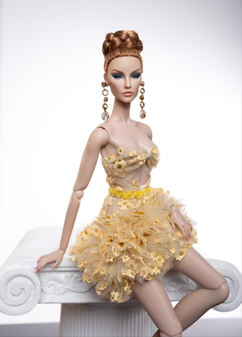 May include: A fashion doll with long, auburn hair styled in a bun, wearing a yellow, floral-patterned dress with a ruffled skirt. The doll is sitting on a white pedestal.