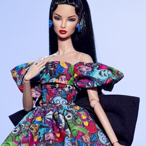May include: A fashion doll with long black hair, red lipstick, and blue earrings and bow. The doll wears a colorful dress with a black bow in the back. The dress features a print of cartoon characters in various colors. The doll is posed against a light blue background.