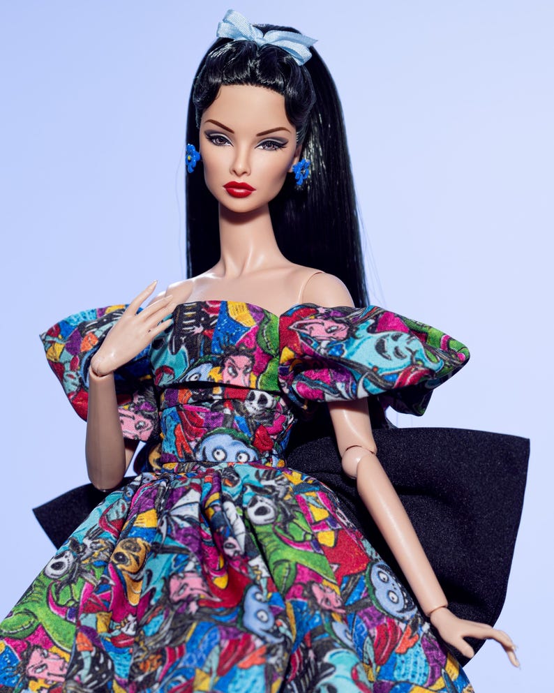 May include: A fashion doll with long black hair, red lipstick, and blue earrings and bow. The doll wears a colorful dress with a black bow in the back. The dress features a print of cartoon characters in various colors. The doll is posed against a light blue background.