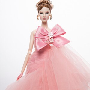 May include: A pink tulle gown with a large pink satin bow and white flower embellishments. The doll is wearing a floral crown and gold earrings.